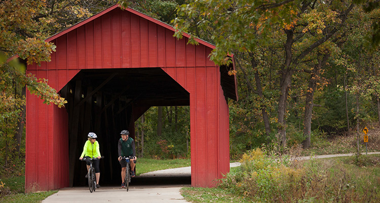 two bikers going through a red covered bridge